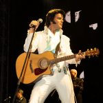 Elvis impersonator JD King in white jumpsuit with stage background